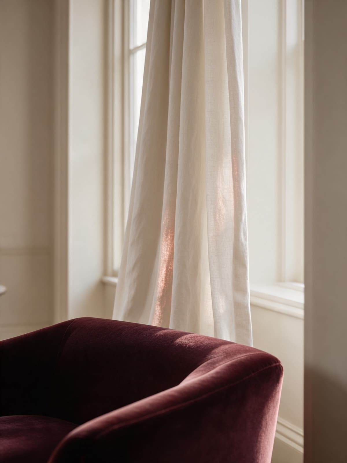 An ivory linen curtain beside a tall window, with a deep maroon velvet armchair in the foreground